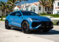 A matte blue Lamborghini Urus SE is parked on a sunny street lined with palm trees and upscale houses, showcasing the luxury of Lamborghini Palm Beach—imagine arriving home in style with a 12-Month Lease.
