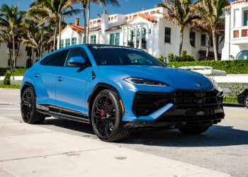 A matte blue Lamborghini Urus SE is parked on a sunny street lined with palm trees and upscale houses, showcasing the luxury of Lamborghini Palm Beach—imagine arriving home in style with a 12-Month Lease.