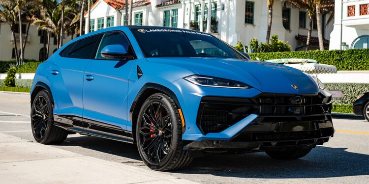 A matte blue Lamborghini Urus SE is parked on a sunny street lined with palm trees and upscale houses, showcasing the luxury of Lamborghini Palm Beach—imagine arriving home in style with a 12-Month Lease.