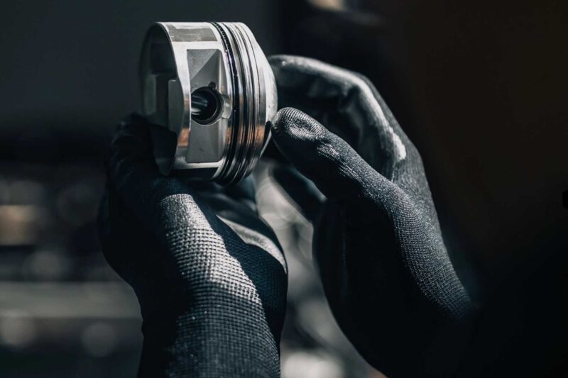 A person wearing black gloves holds and inspects a shiny metal piston, likely for a Lamborghini engine, against a blurred background.