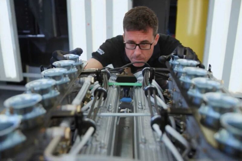 A man wearing glasses and black gloves inspects a large, complex Lamborghini engine with multiple cylinders in a well-lit workshop, capturing the spirit of Polo Storico heritage.