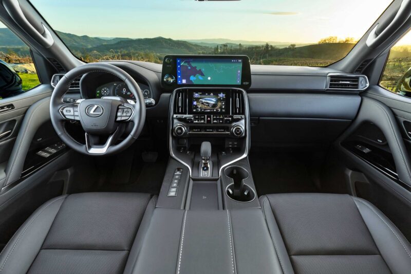 Interior view of the 2025 Lexus LX 700h Overtrail 4WD showcases its steering wheel, dashboard, touchscreen displays, and front seats—all set against a scenic landscape. Review: Highly capable and moderately practical for luxury SUV enthusiasts.
