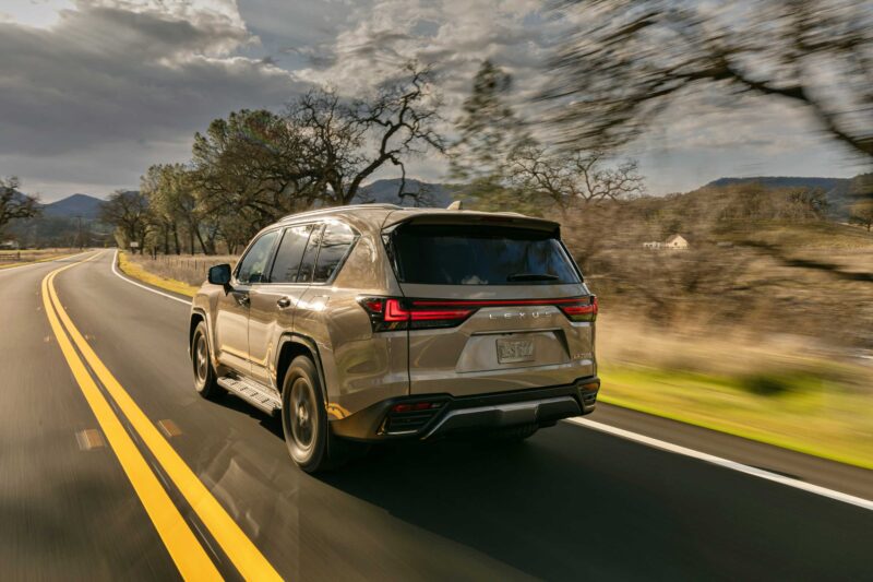A beige SUV, much like the 2025 Lexus LX 700h Overtrail 4WD, drives down a two-lane road bordered by grass and leafless trees under a cloudy sky.