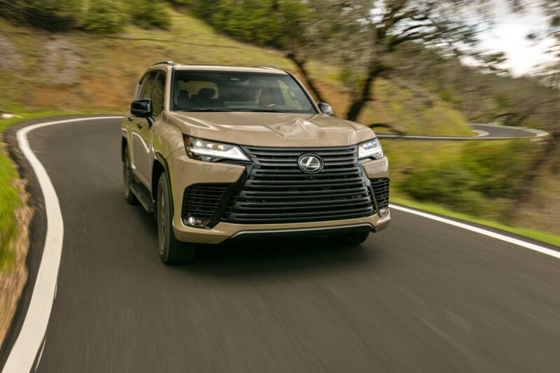 A tan Lexus SUV, likely the highly capable 2025 Lexus LX 700h Overtrail 4WD, drives on a winding, two-lane road through a green, wooded area during the day.