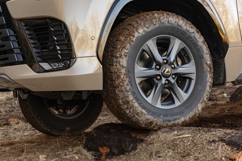 Close-up of a Lexus SUV front wheel with mud-covered tire driving over a rock on a dirt surface. Review: 2025 Lexus LX 700h Overtrail 4WD is highly capable and moderately practical in tough off-road conditions.