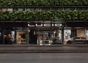 Street view of a Lucid car showroom with large windows, a visible Lucid electric car inside, and green foliage covering the upper facade of the building.