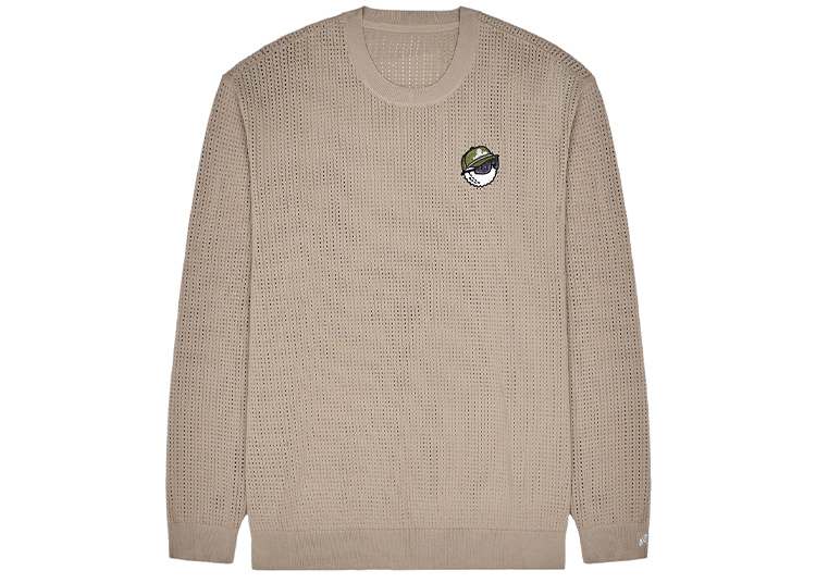 A beige, long-sleeve knit sweater with a round neck from the Malbon Golf Capsule Collection, featuring a small embroidered patch of a cartoon face wearing a green cap on the left chest.