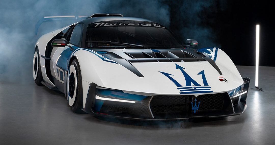 A white and black Maserati race car with blue accents and the Maserati trident logo on the hood, displayed indoors with smoke in the background.