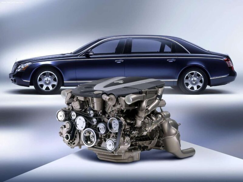 A luxury sedan is parked in the background with a detailed V12 engine displayed prominently in the foreground, showcasing how V12 Lives On: Mercedes-Benz Will Retain the Flagship 12-Cylinder Into The 2030s.