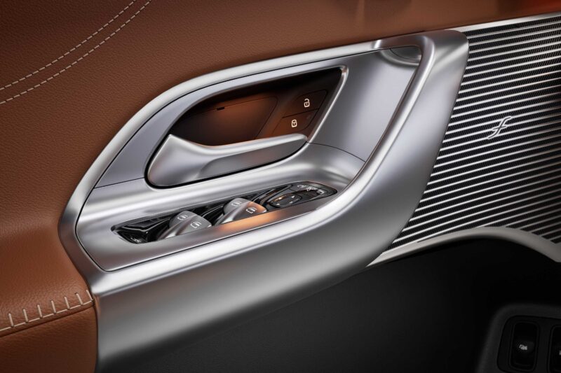 Close-up of a Mercedes car door interior showing window and lock controls, metallic trim, brown leather upholstery, and a stitched seam—crafted to complement innovations like the 39-Inch Hyperscreen found in the All-Electric GLC.
