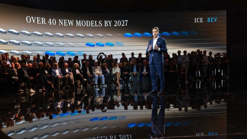A man in a suit presents on stage to an audience, with a screen behind him displaying "OVER 40 NEW MODELS BY 2027" and graphics labeled ICE and BEV at Beyond the All-electric GLC: Mercedes-Benz at IAA 2025.