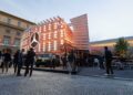 People gather around a modern Mercedes-Benz display pavilion with illuminated logo in a city square, featuring a green vehicle and outdoor seating area—showcasing Beyond the All-electric GLC: Mercedes-Benz at IAA 2025.