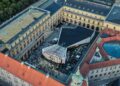 Aerial view of a large, historic building complex with a modern, angular pavilion in the central courtyard, surrounded by people and outdoor seating areas—perfect for experiencing Beyond the All-electric GLC: Mercedes-Benz at IAA 2025.