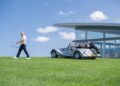 A man walks on a grassy lawn holding a golf club, with a Morgan Plus Four convertible and golf bag parked nearby in front of a modern glass building, marking its East Coast debut under the blue Long Island sky.