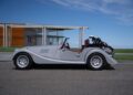 A silver Morgan Plus Four convertible sports car parked on a road, with a modern building and green lawn in the background under a blue sky—capturing its East Coast Debut with our Long Island Partner.