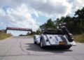 A silver Morgan Plus Four convertible with a golf bag attached drives up a road toward a “Chevron Gasolines” sign, with trees and clouds in the background, marking its East Coast debut.