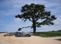 A vintage Morgan Plus Four convertible is parked on a sandy area near a large tree, with a grassy lawn in the background beneath a blue sky—capturing the charm of its East Coast debut with a Long Island partner.