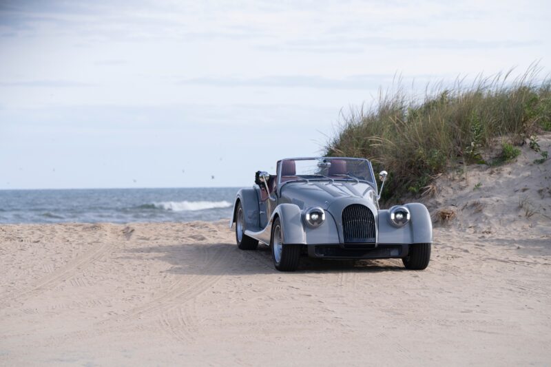 A classic Morgan Plus Four convertible is parked on sandy terrain near grass-covered dunes, with the ocean in the background, capturing the spirit of its East Coast debut on Long Island.