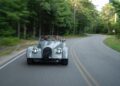 A silver Morgan Plus Four vintage-style convertible glides along a winding, tree-lined road on Long Island, with a man behind the wheel.