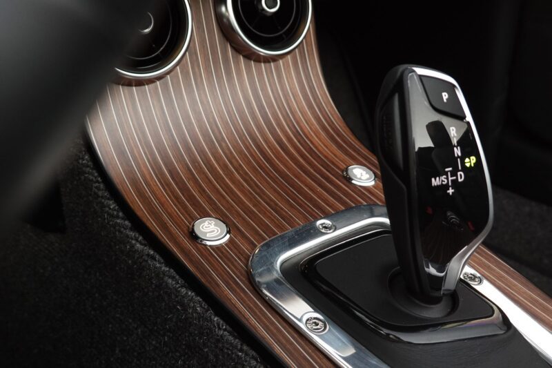 Close-up of a car’s automatic gear shift lever on a wood-patterned center console, echoing the refined craftsmanship seen in the 2025 Morgan Plus Four Review: A Timeless Reminder of Olde-School Driving Fun.