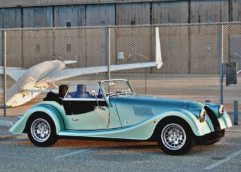 A classic convertible sports car with a teal and black finish, reminiscent of the 2025 Morgan Plus Four Review: A Timeless Reminder of Olde-School Driving Fun, is parked in front of a chain-link fence and a small airplane at an airport.