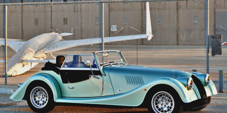 A classic convertible sports car with a teal and black finish, reminiscent of the 2025 Morgan Plus Four Review: A Timeless Reminder of Olde-School Driving Fun, is parked in front of a chain-link fence and a small airplane at an airport.