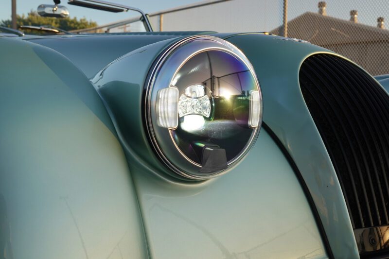 Close-up of a round headlight on a vintage turquoise car, echoing the classic appeal found in the 2025 Morgan Plus Four review: a timeless reminder of olde-school driving fun, with reflections dancing on glass and across its curved hood and grille.