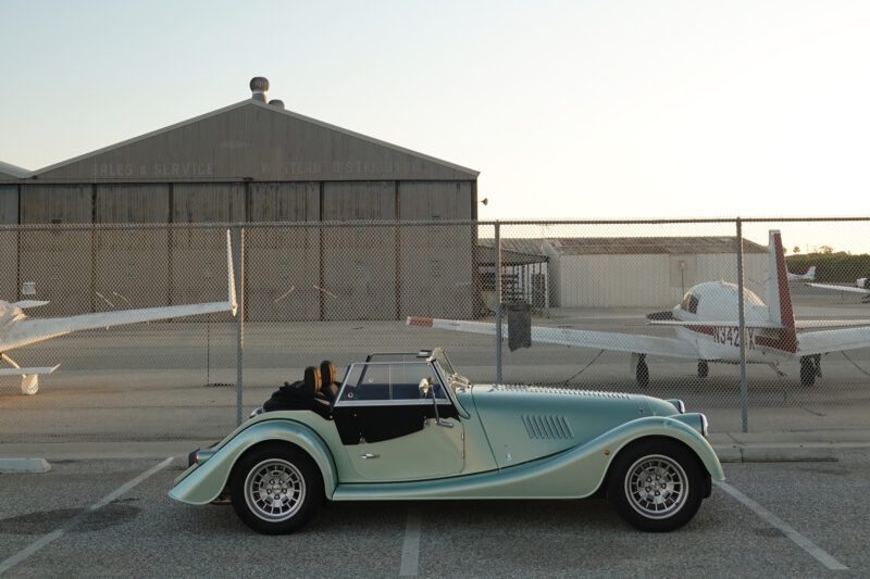 A vintage convertible car, reminiscent of the 2025 Morgan Plus Four Review: A Timeless Reminder of Olde-School Driving Fun, is parked in front of a chain-link fence with two small airplanes and a hangar in the background.