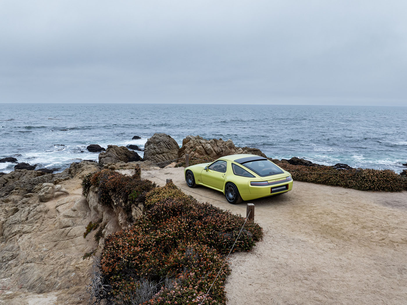 A yellow Nardone 928 production car is parked on a dirt path overlooking rocky cliffs and the ocean under a cloudy sky.