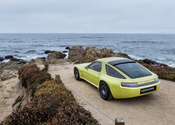 A yellow Nardone 928 sports car is parked on a dirt path overlooking rocky cliffs and the ocean under a cloudy sky.