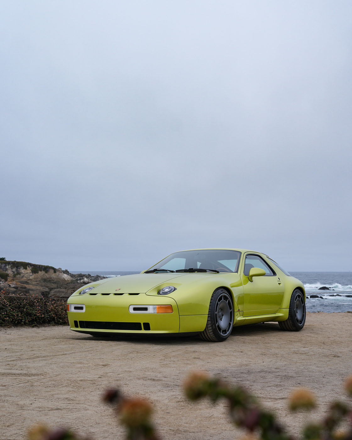 A light green Nardone 928 sports car parked on a sandy area near the ocean, with cloudy skies and a rocky coastline in the background.