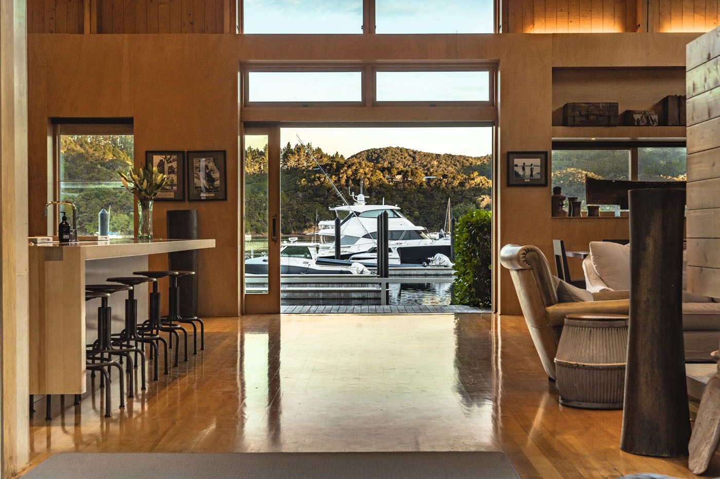 Modern open-plan living space with wooden floors, a kitchen island with barstools, and large windows overlooking a dock with boats and the scenic New Zealand coastline framed by a tree-covered hillside—perfect for waterfront ownership.