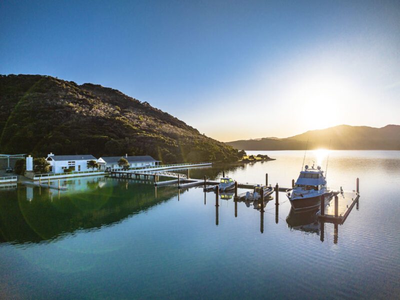 A boat docked at a pier on calm water along the coveted coastline, with surrounding hills and buildings illuminated by the rising or setting sun.