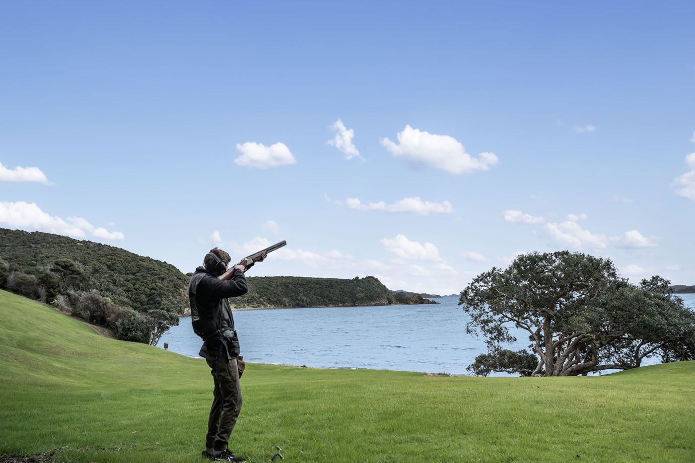 A person stands on green grass near water along New Zealand’s coveted coastline, aiming a shotgun at the sky under a mostly clear, blue sky with scattered clouds.