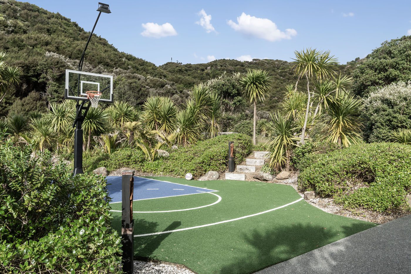 Outdoor half basketball court with a blue and green surface, nestled among lush greenery, palm trees, and hills under a partly cloudy sky—your own slice of coveted coastline reminiscent of the breathtaking New Zealand coastline.