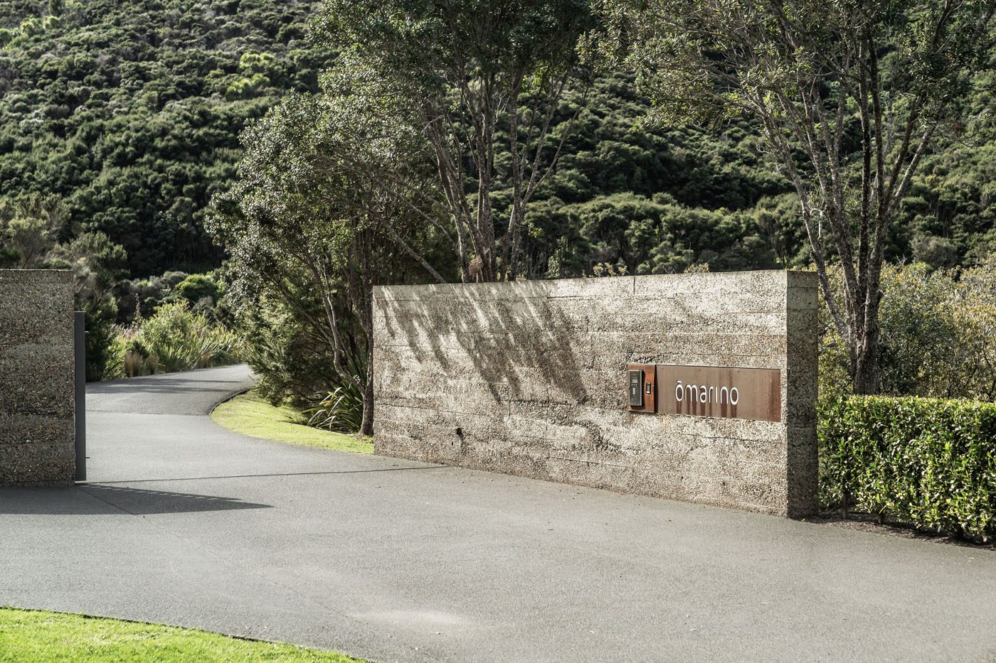 A paved driveway curves past a textured concrete wall with a sign that reads "d/manino," set against dense green trees and bushes, ideally situated along New Zealand's coveted coastline for true ownership.