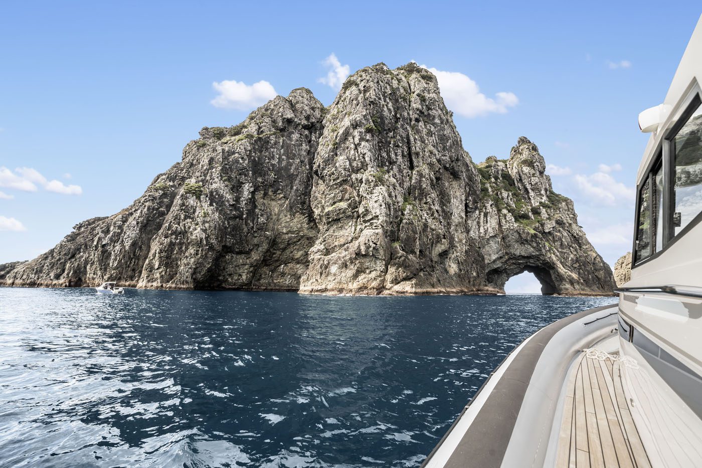 A rocky island with a large arch formation rises from the ocean along the coveted New Zealand coastline, viewed from a boat on calm blue water under a clear sky.