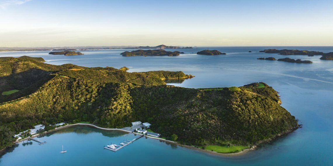 Aerial view of a lush, green peninsula along the coveted coastline, surrounded by calm blue water, with docks, buildings, and small islands visible in the distance under a clear sky.