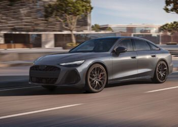 A sleek gray 2026 Audi A6 with black wheels glides down a city street beside modern buildings and trees, under daylight.