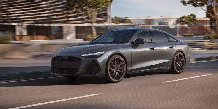 A sleek gray 2026 Audi A6 with black wheels glides down a city street beside modern buildings and trees, under daylight.