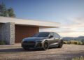 A gray 2026 Audi A6 luxury sedan is parked in front of a modern house with stone and wood accents, set against a scenic, partly cloudy sky.