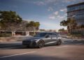 A sleek, dark-colored 2026 Audi A6 glides along a city street lined with modern buildings and trees under a partly cloudy sky, perfectly embodying sophistication at an attractive Audi A6 price.