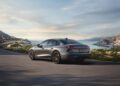 A silver 2026 Audi A6 sedan drives on a winding coastal road with mountains, rocks, and water in the background under a partly cloudy sky.