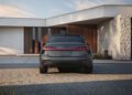 A matte gray 2026 Audi A6 sedan is parked in front of a modern house with stone and wood exterior details, viewed from the rear—perfect for those curious about the new Audi A6 price.