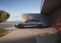A sleek grey 2026 Audi A6 is parked next to a modern stone and wood house, with trees and mountains visible in the background under a clear sky.