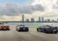 Three sports cars, including a McLaren, drive on a highway bridge with a city skyline and waterfront in the background under a cloudy sky—capturing the thrill of a road trip and the spirit of States of Endurance.