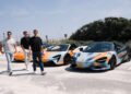 Four men walk past three parked McLaren sports cars in a sunny outdoor parking lot, suggesting the start of an epic road trip under a blue sky with lush greenery—a true scene of States of Endurance.