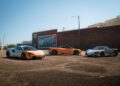 Three sports cars, including a sleek McLaren, are parked in a mostly empty outdoor gravel lot near a brick building with a mural—an ideal stop during a States of Endurance road trip.