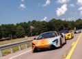 Three brightly-colored sports cars, including a McLaren, drive on a highway lined with trees and a metal guardrail, an American flag waving in the background—a perfect scene from a States of Endurance road trip under a partly cloudy sky.