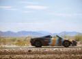 A McLaren sports car with a multicolored paint job is parked on a road in a desert landscape, mountains rising in the distance under a partly cloudy sky—ready for an epic States of Endurance road trip.